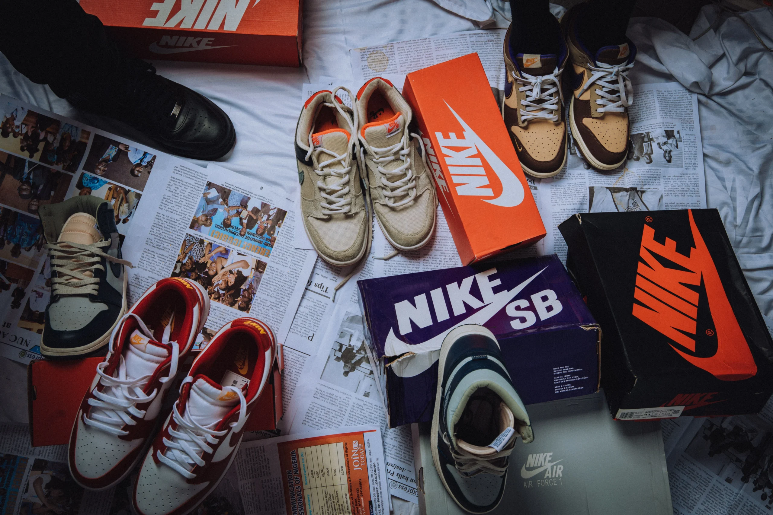 Collection of Nike trainers and shoeboxes arranged across magazines and newspapers on a bed, showcasing different sneaker styles in an editorial flat lay that reflects sneaker collecting and streetwear culture.