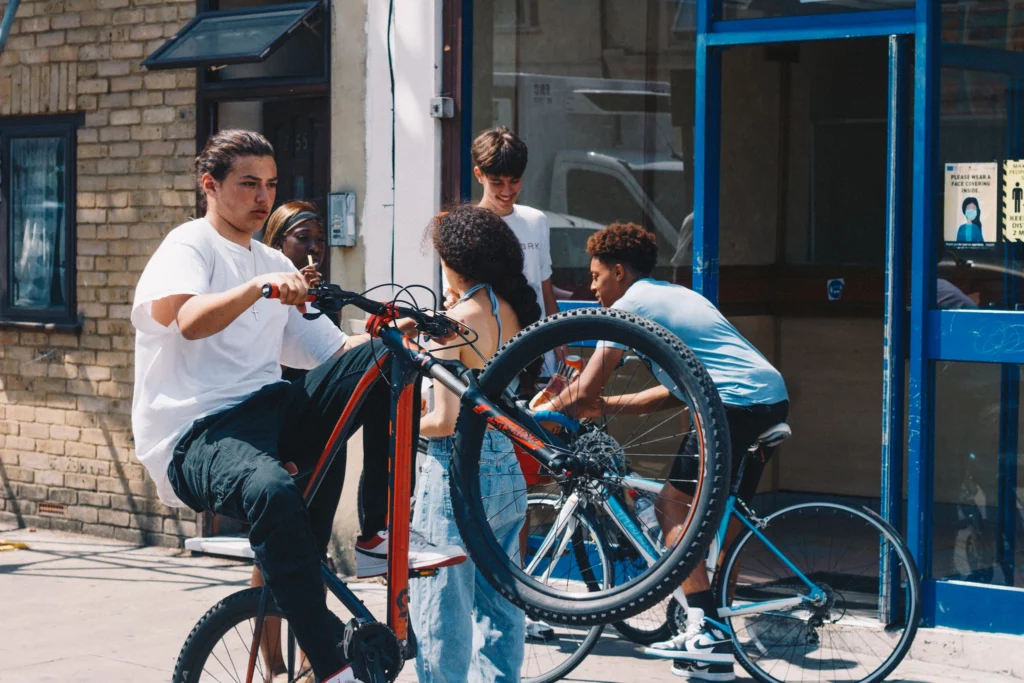 Youth Subcultures: What They Are and How to Identify Them Teenager pulling a wheelie on a mountain bike outside a shopfront while friends gather with their bikes, showing a craft-led youth subculture